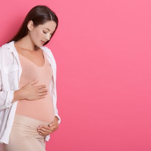 Beautiful pregnant woman hugging her tummy against pink background, looking at her belly with love, copy space for advertisement or promotional text text.